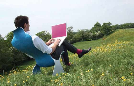 Man sitting in a modernist chair in a field of buttercups pretending to type on a laptop with a blank pink screen